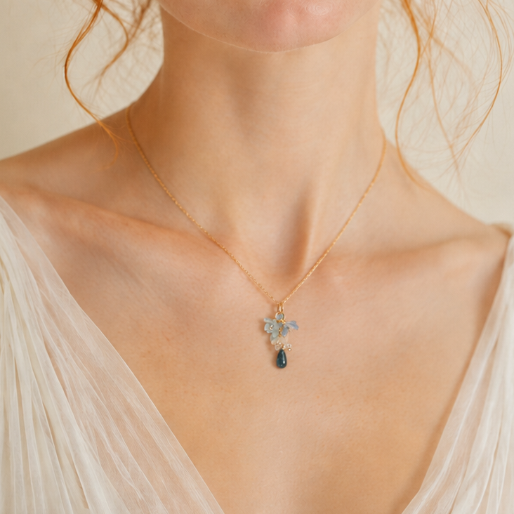 A close-up portrait of a woman with red hair wearing a delicate gold necklace with a small silver-toned cross pendant featuring a dark blue stone, set against a soft beige background.