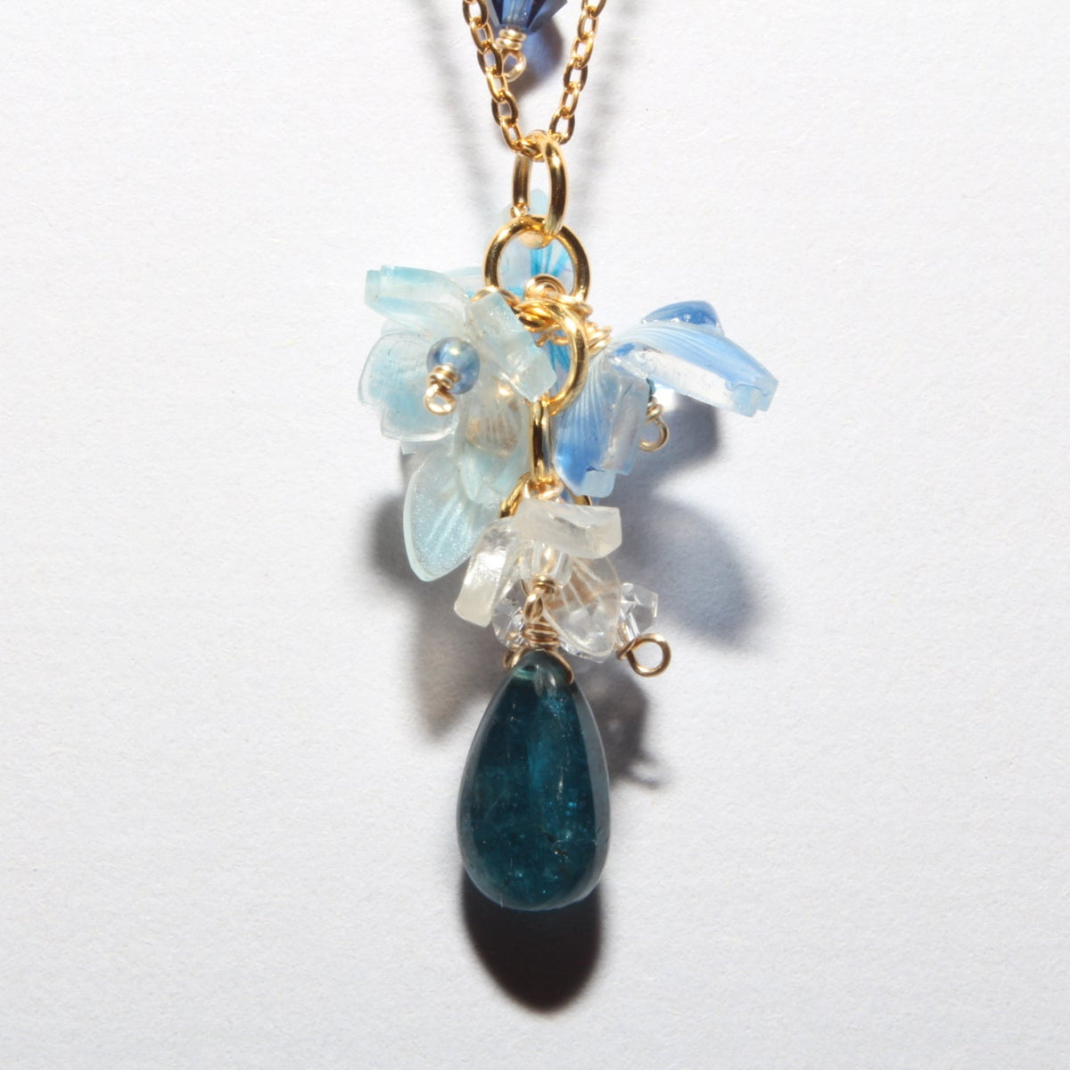 A delicate gold chain necklace featuring a floral pendant with light blue and clear crystals, accented by a dark blue teardrop stone at the bottom, shown against a plain white background.