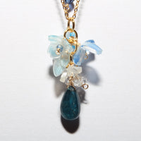 A delicate gold chain necklace featuring a floral pendant with light blue and clear crystals, accented by a dark blue teardrop stone at the bottom, shown against a plain white background.