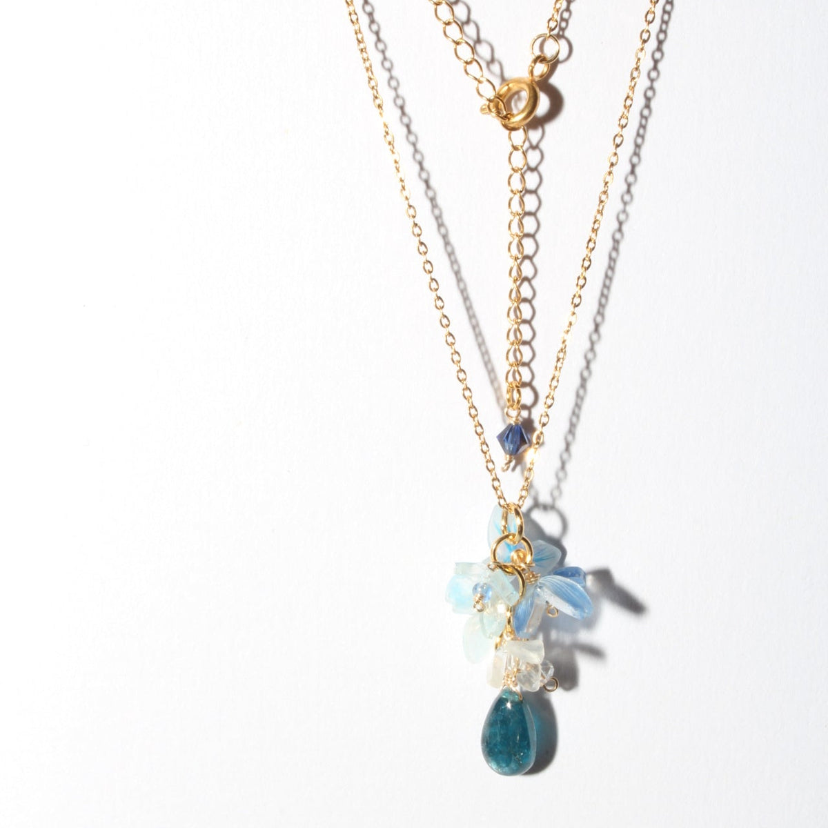 A gold layered necklace with a floral and teardrop pendant, featuring light blue and white flowers, a small dark blue bead, and a larger blue teardrop stone, shown on a plain white background.