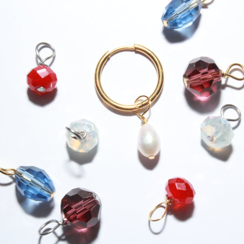 A set of gold-toned jewelry findings or beaded components arranged on a white background, including a circular gold ring, a pearl-like teardrop bead, faceted beads in red, blue, and clear, and small metal loops.