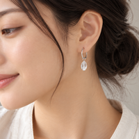 A close-up of a woman wearing a silver huggie hoop earring with a clear teardrop-shaped crystal pendant.
