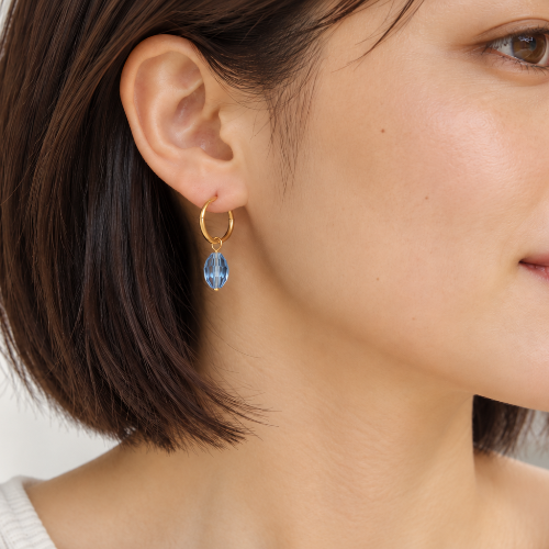 A close-up side profile of a woman wearing a delicate gold hoop earring with a teardrop-shaped light blue gemstone dangle.