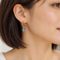 A close-up side profile of a woman wearing a delicate gold hoop earring with a teardrop-shaped light blue gemstone dangle.