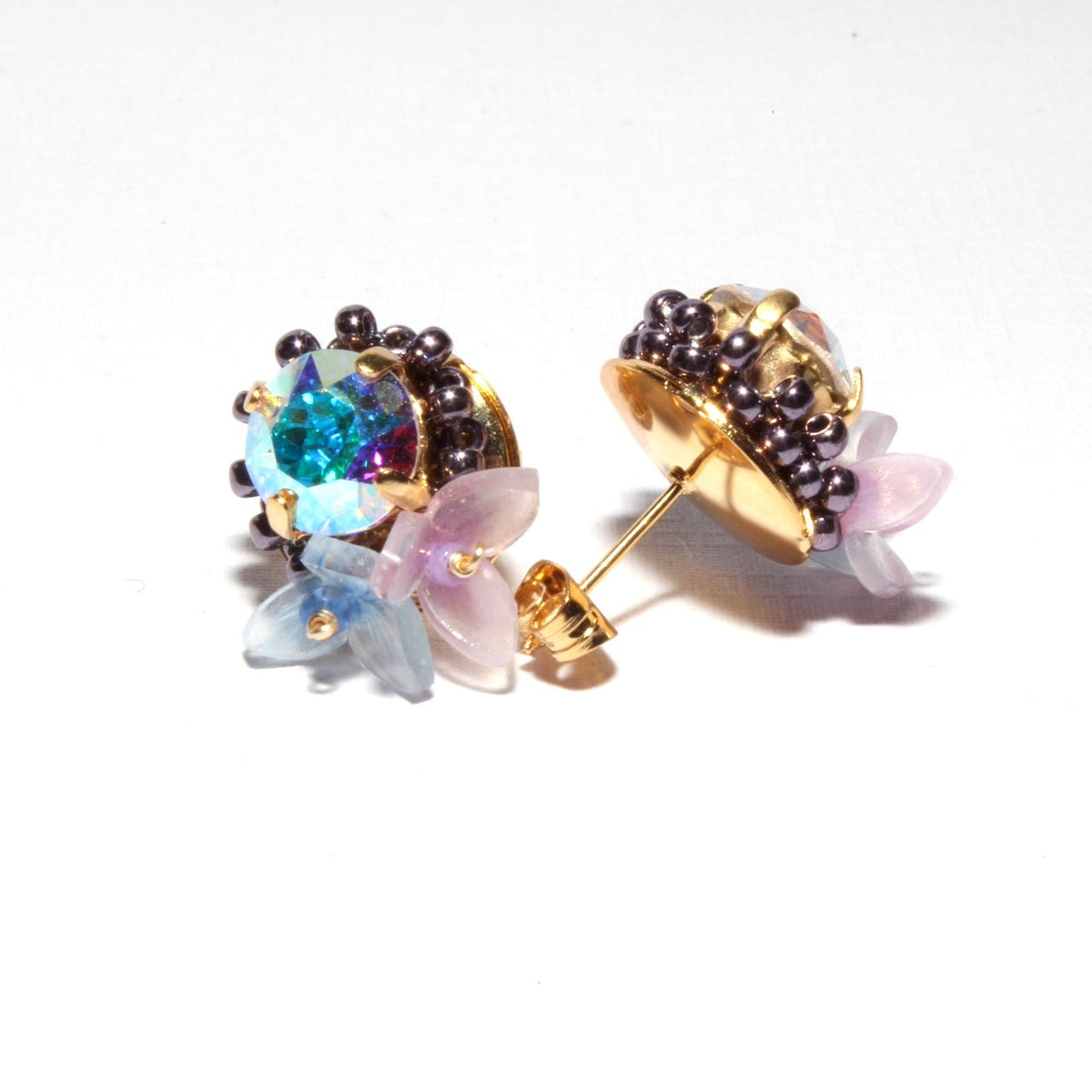 A pair of gold stud earrings featuring a central iridescent blue gemstone, surrounded by small dark purple beads and delicate translucent pink and pale blue flower petals, shown on a plain white background.