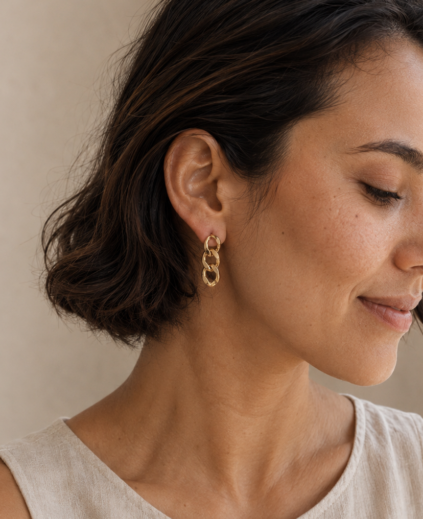 A woman with short dark brown hair wearing a pair of gold chain-link drop earrings and a light beige linen top, shown in a close-up profile against a soft beige background.