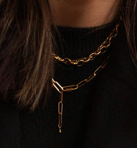 A close-up of a gold paperclip chain earring and matching chain necklace worn by a model in a black top, with brown hair framing the side of the face.