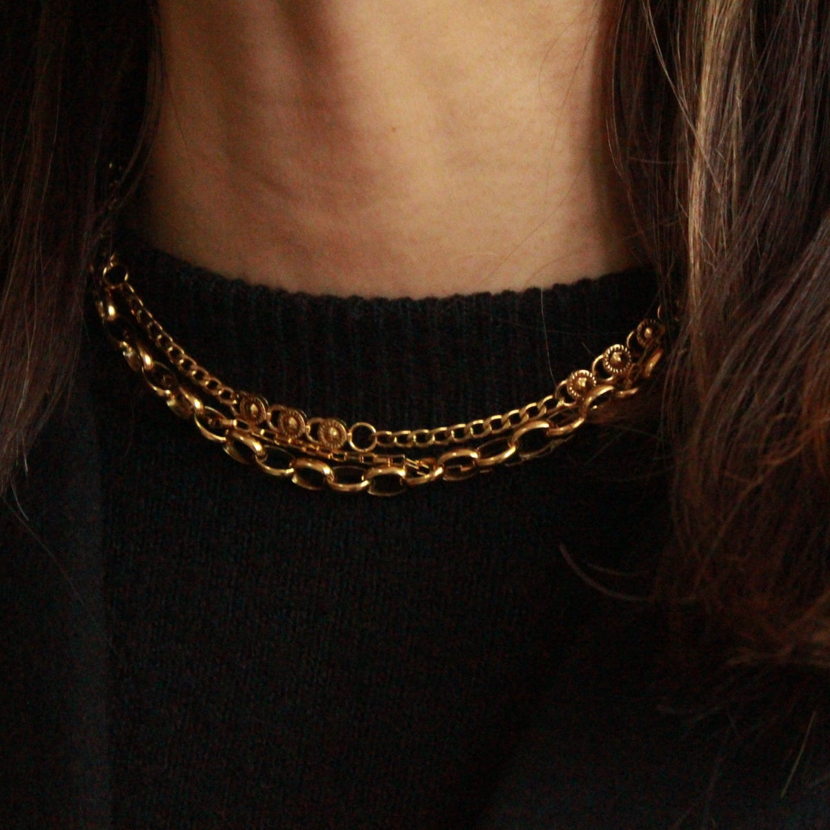 A close-up of a person wearing a double-layered gold chain necklace over a black ribbed sweater, with dark brown hair framing the neck.