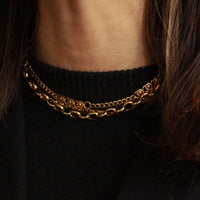 A close-up of a person wearing a double-layered gold chain necklace over a black ribbed sweater, with dark brown hair framing the neck.