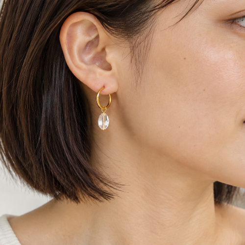 A close-up side profile of a woman wearing a delicate gold hoop earring with a clear teardrop-shaped crystal drop, set against a soft, light gray background.