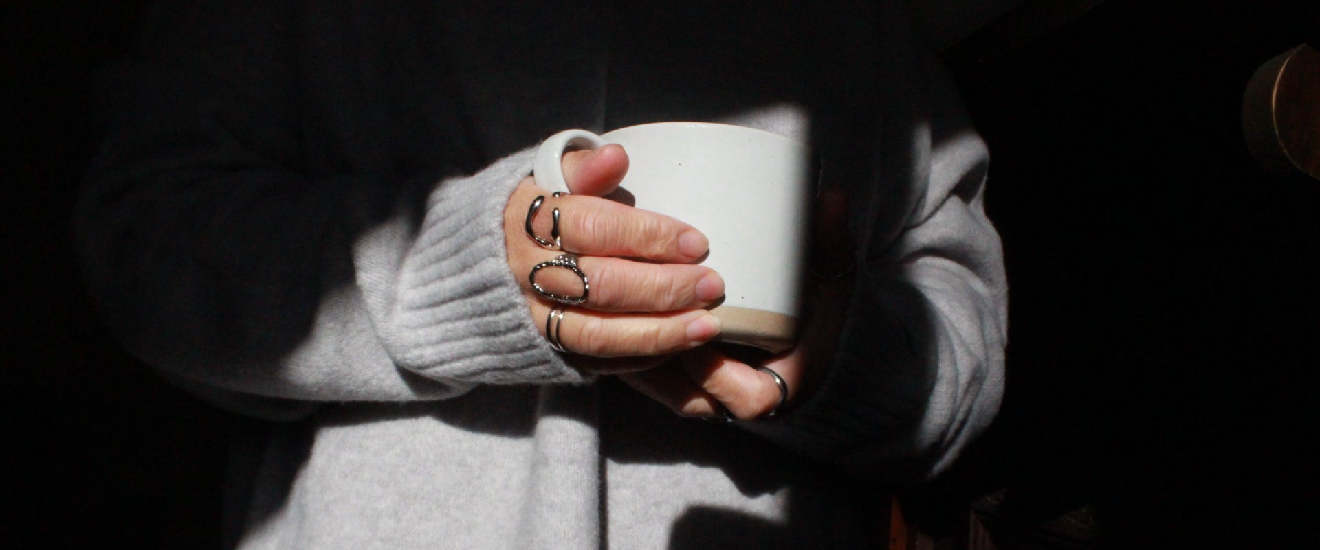A person in a light gray knit sweater holds a white ceramic mug in their hands, which are adorned with multiple stacked rings, set against a dark background with dramatic side lighting.
