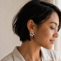 A close-up side profile of a woman with short dark hair wearing a small silver star-shaped stud earring and a light beige collared shirt against a soft beige background.