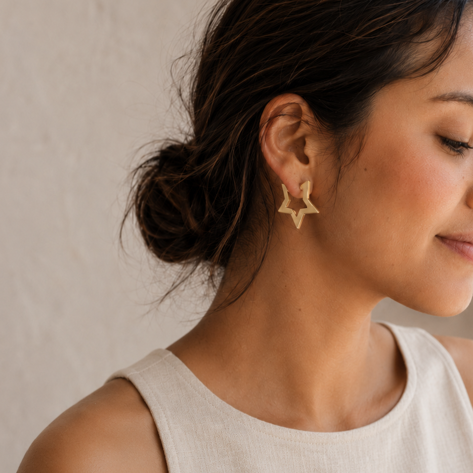 A woman with her hair in a low bun is shown in profile wearing a small gold star-shaped stud earring. She is wearing a light beige sleeveless top against a neutral, textured beige wall.