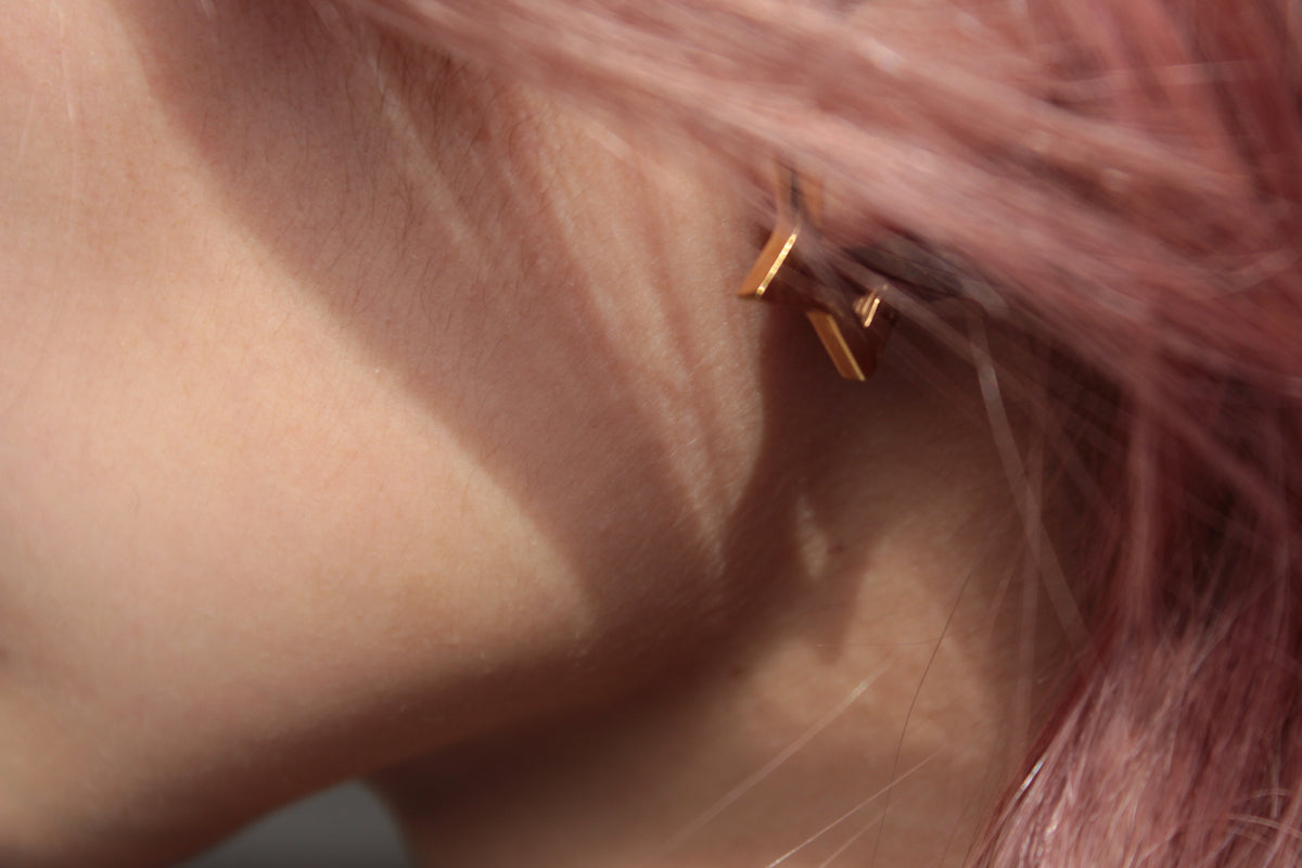 A close-up of a person's ear with a small gold geometric stud earring, framed by pink hair.