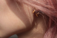 A close-up of a person's ear with a small gold geometric stud earring, framed by pink hair.