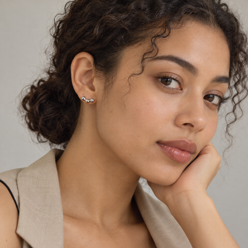 A close-up portrait of a woman with curly brown hair wearing a small silver and black wavy earring, styled with a beige blazer over a black top against a neutral gray background.