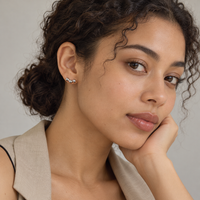 A close-up portrait of a woman with curly brown hair wearing a small silver and black wavy earring, styled with a beige blazer over a black top against a neutral gray background.