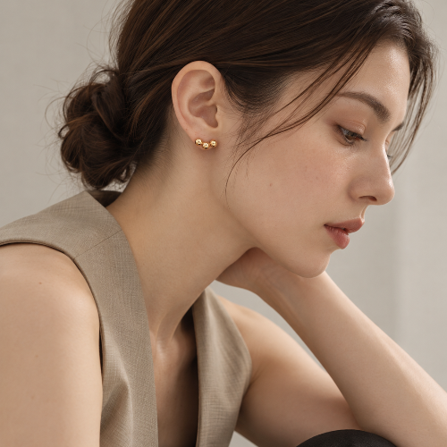 A close-up of a woman wearing a small gold stud earring with three tiny gold triple balls styled with a beige sleeveless vest and black pants against a neutral gray background.