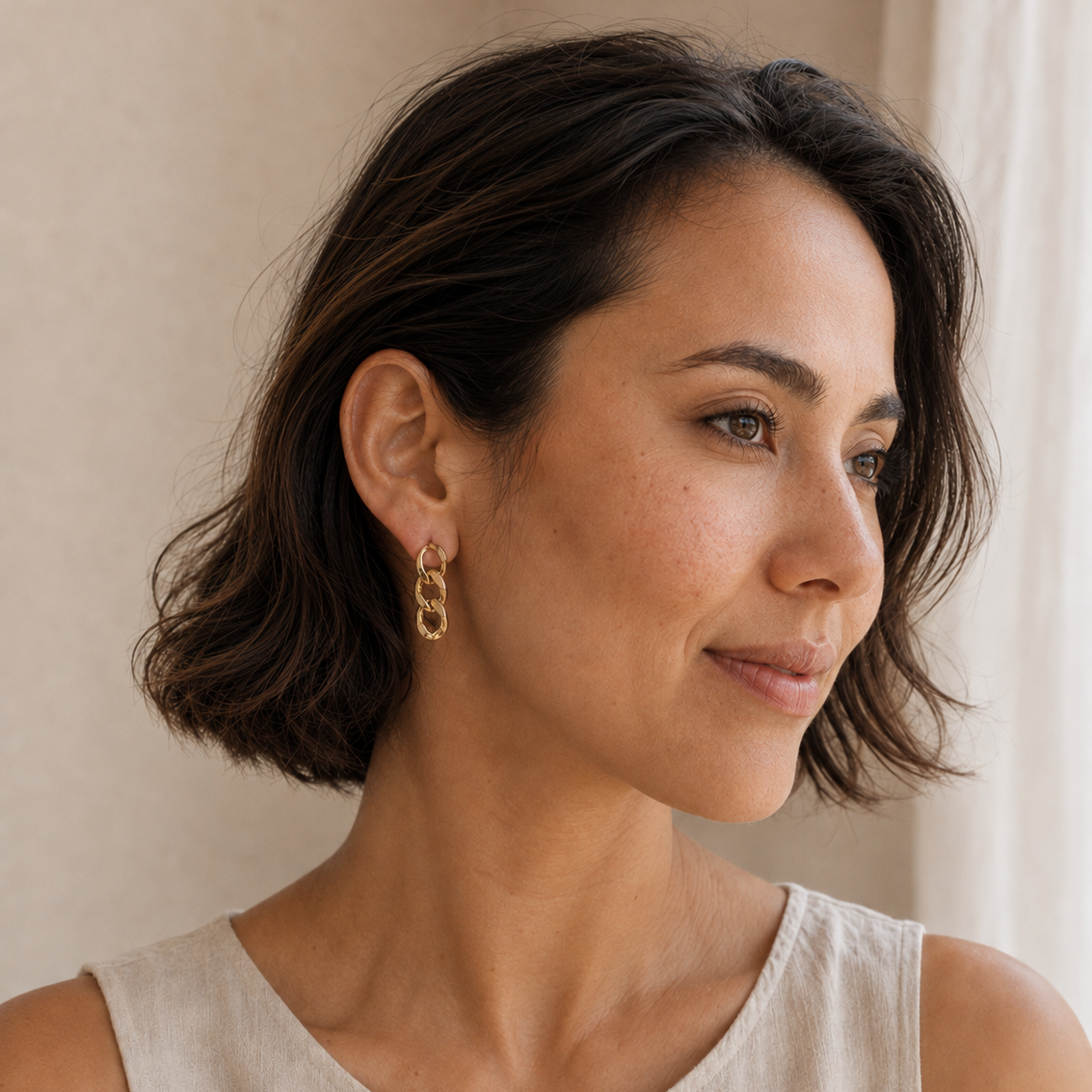 A woman with short dark brown hair wearing a pair of gold chain-link drop earrings and a light beige linen top, shown in a close-up profile portrait against a soft beige background.