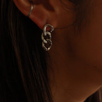 A close-up of a person's ear wearing a silver hoop earring with a dangling interlocking chain-link drop earring, against dark hair and warm lighting.