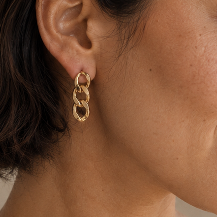 A woman with short dark brown hair wearing a pair of gold chain-link drop earrings and a light beige linen top, shown in a close-up profile against a soft beige background.
