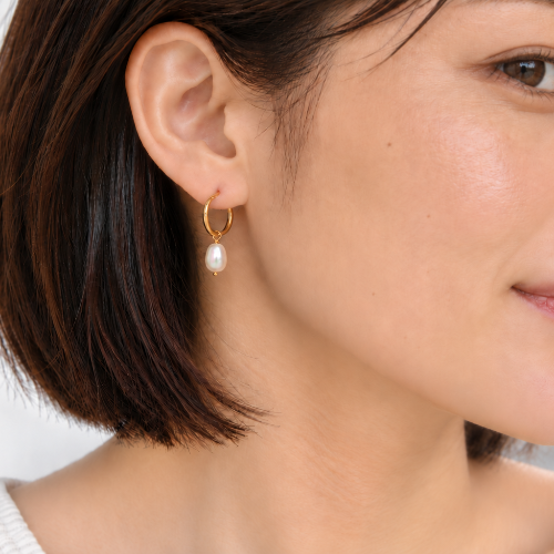 A close-up of a woman wearing a delicate gold hoop earring with a single white pearl drop, shown against a soft light gray background.