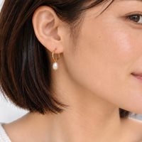A close-up of a woman wearing a delicate gold hoop earring with a single white pearl drop, shown against a soft light gray background.
