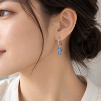 A close-up of a woman wearing a silver huggie hoop earring with a dangling light blue teardrop gemstone, shown in profile against a soft neutral background.