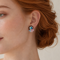 A close-up of a woman with red hair wearing a delicate floral stud earring with a blue center and pale pink petals, set against a soft beige background.