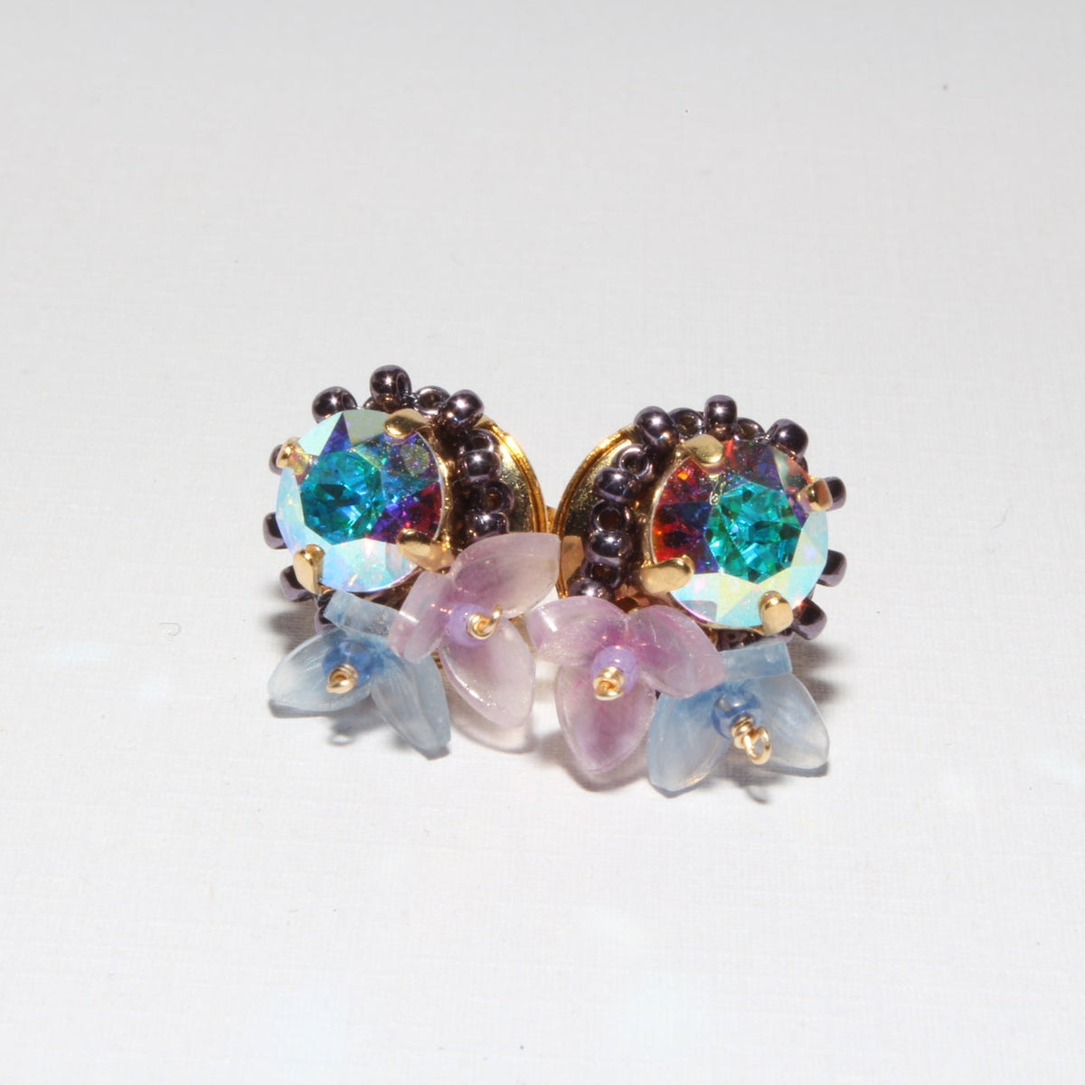 A pair of gold-toned stud earrings featuring iridescent blue-green teardrop crystals, surrounded by dark brown beads and decorated with translucent pastel pink and blue flower petals, set against a plain white background.