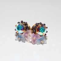 A pair of gold-toned stud earrings featuring iridescent blue-green teardrop crystals, surrounded by dark brown beads and decorated with translucent pastel pink and blue flower petals, set against a plain white background.