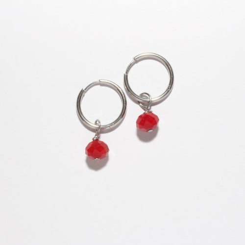 A pair of silver-toned huggie hoop earrings with a small red bead dangle, shown on a plain white background.