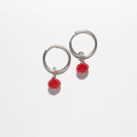 A pair of silver-toned huggie hoop earrings with a small red bead dangle, shown on a plain white background.