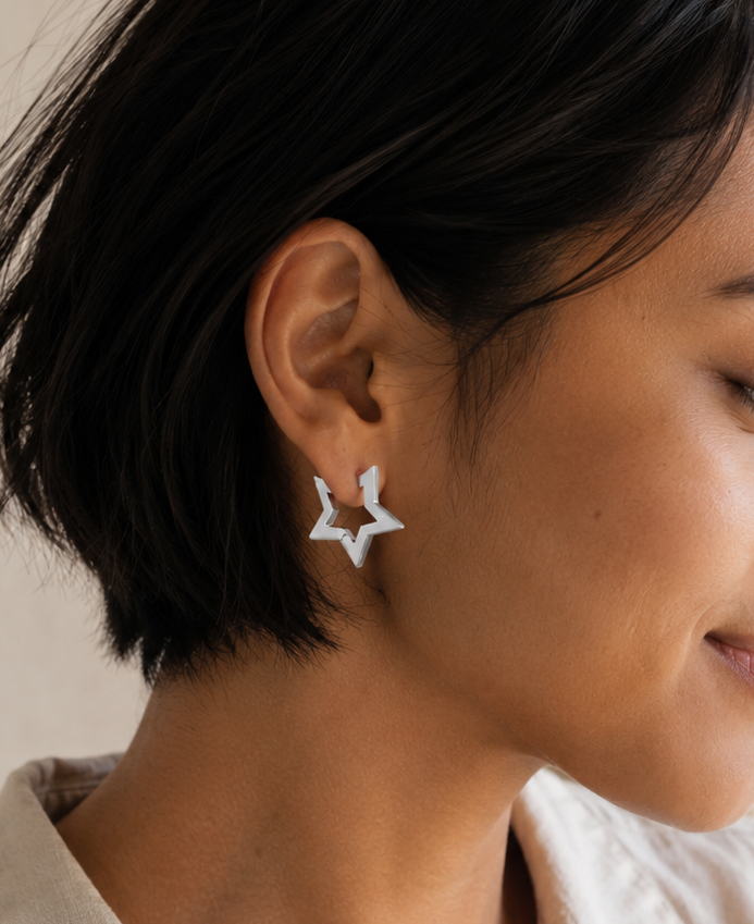 A close-up side profile of a woman with short dark hair wearing a small silver star-shaped stud earring and a light beige collared shirt against a soft beige background.