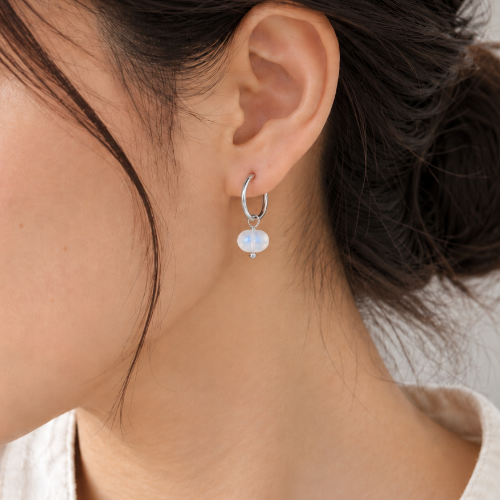 A close-up of a woman wearing a delicate silver huggie hoop earring with a small white pearl drop, shown from the side.